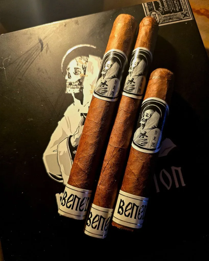 Three BLTC Benediction cigars in Robusto, Lonsdale, and Toro vitolas resting on the Benediction black box artwork under dramatic warm lighting at Smokers Abbey Austin