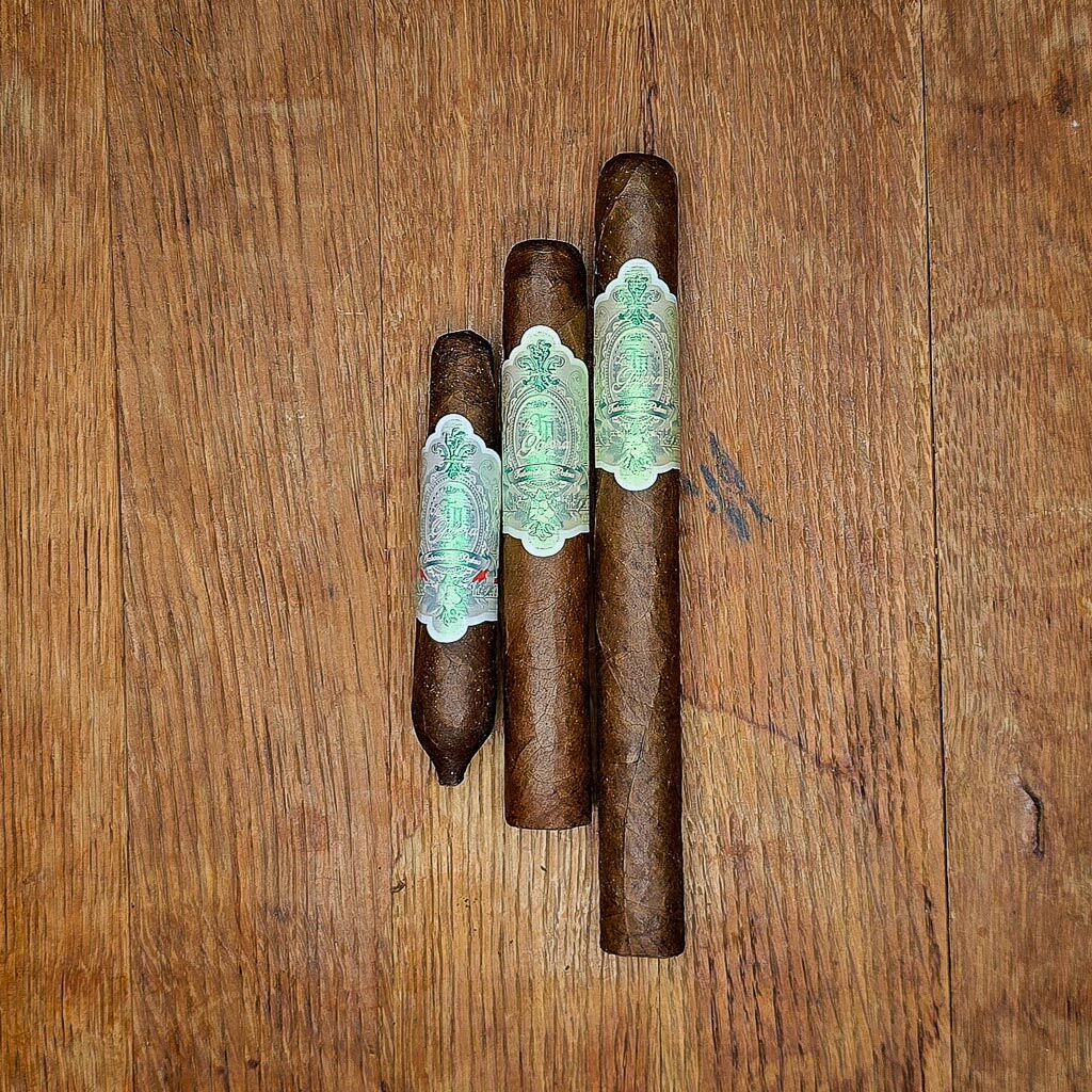Three La Galera Imperial Jade cigars in Chiquito Perfecto, Robusto, and Churchill vitolas displayed side by side on rustic wood surface showing dark African Cameroon wrapper and iridescent green band at Smokers Abbey Austin