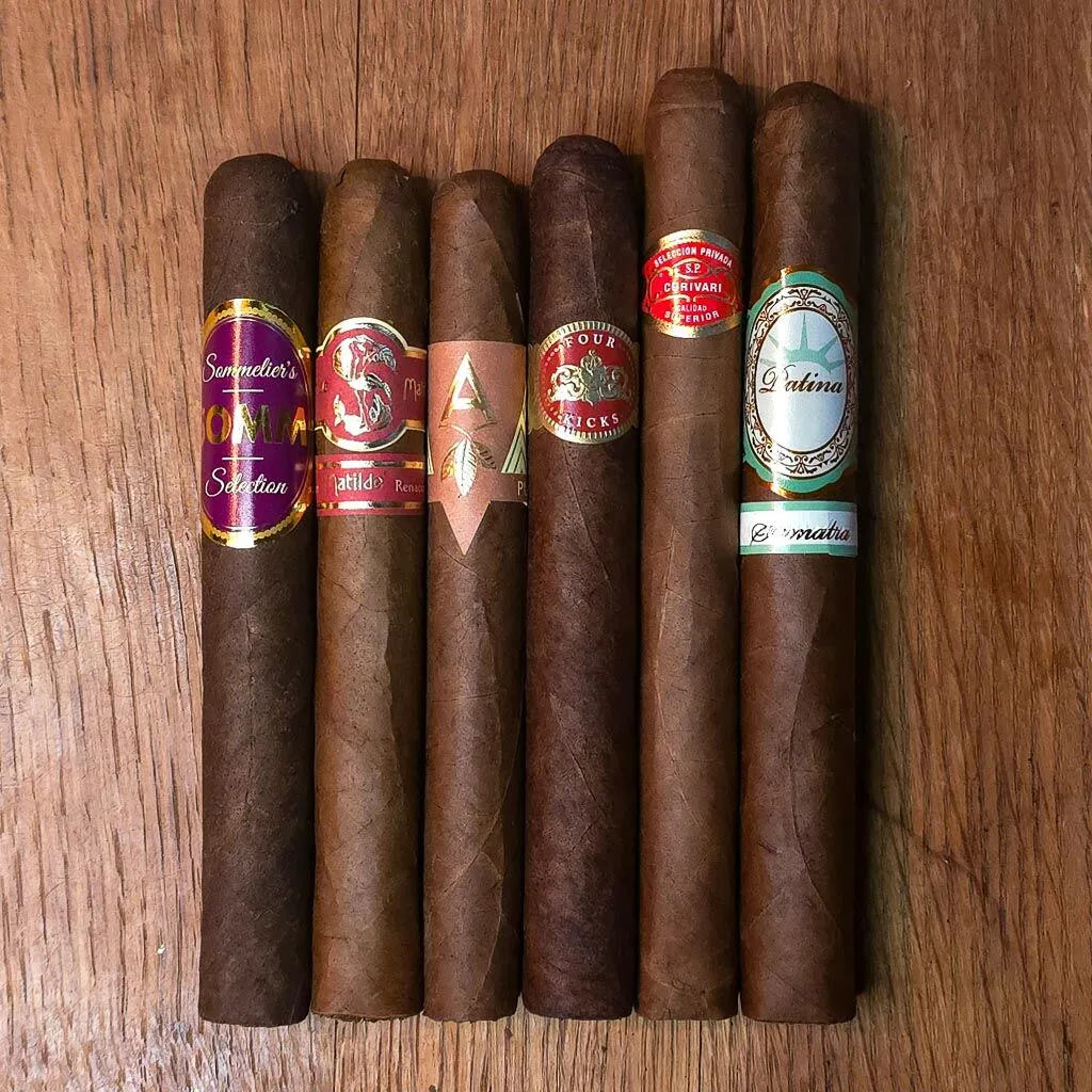 Corona cigar sampler featuring Somm BDX, Matilde Renancer, Artista Puro Ambar, Crowned Heads Four Kicks, Curivari Seleccion Privada, and Patina Sumatra Copper coronas displayed side by side on rustic wood surface at Smokers Abbey Austin