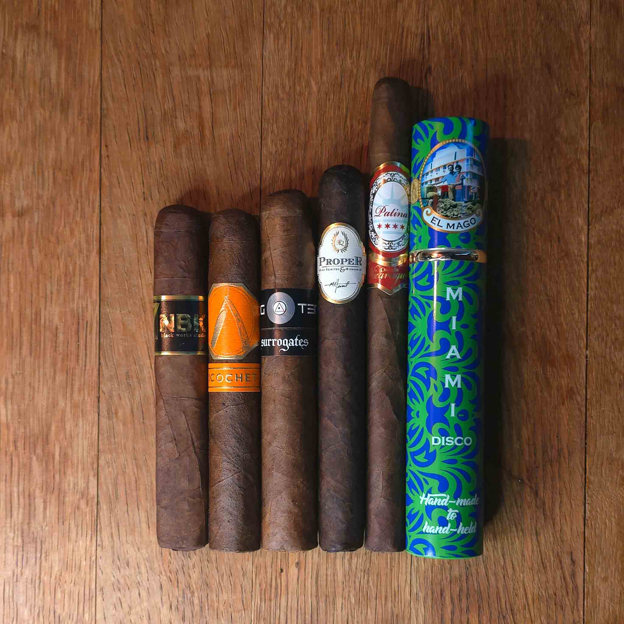 Oscuro Sampler featuring BLK WKS Studio NBK, La Barba Ricochet Crü Oscuro, L'Atelier Surrogates Big Ten, Proper by FQ Cigars, Patina Oro de Nicaragua Lancero, and El Mago Disco displayed side by side on rustic wood surface at Smokers Abbey Austin