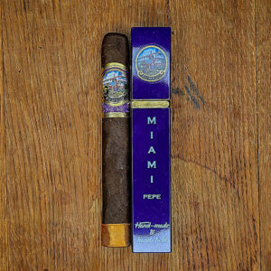 An El Mago Pepe box-pressed Toro cigar displayed on a wooden surface beside its purple Miami Pepe-branded cigar tube.