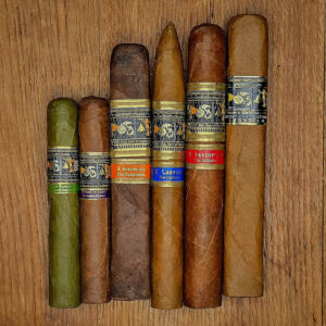 A lineup of six Apostate cigars arranged on a wooden surface, including the Secret Combinations limited release, The Initiatory, The Liahona, The Zarahelma, The Feathered Serpent, and The Sword of Laban.