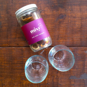 A Mixy Red Wine Sangria cocktail kit jar lying on a wooden surface beside two clear stemless wine glasses.