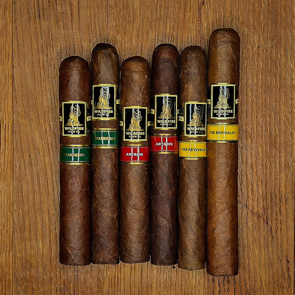 A lineup of six Wildfire Cigars arranged on a wooden surface, featuring two vitolas each of The Wanderer, Artaios, and The Revivalist.