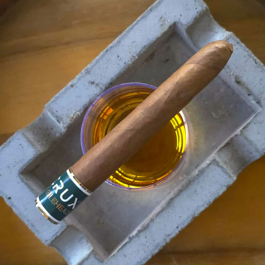 Top-down view of a Crux Marblehead cigar showing the rounded 109 cap positioned over a glass of bourbon.