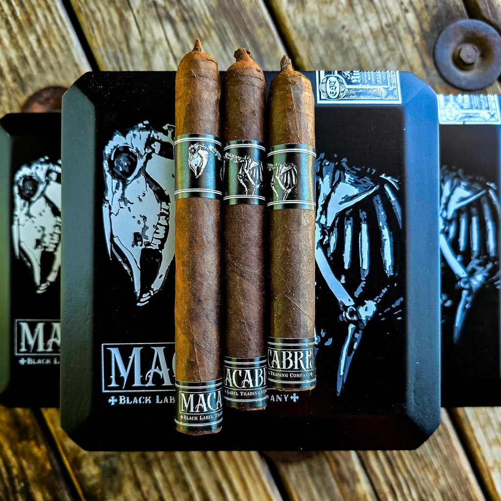 Three BLTC Macabre cigars in Robusto, Corona Gorda, and Toro vitolas standing upright on the black Macabre box with skull artwork — PCA 2026 exclusive limited release at Smokers Abbey Austin