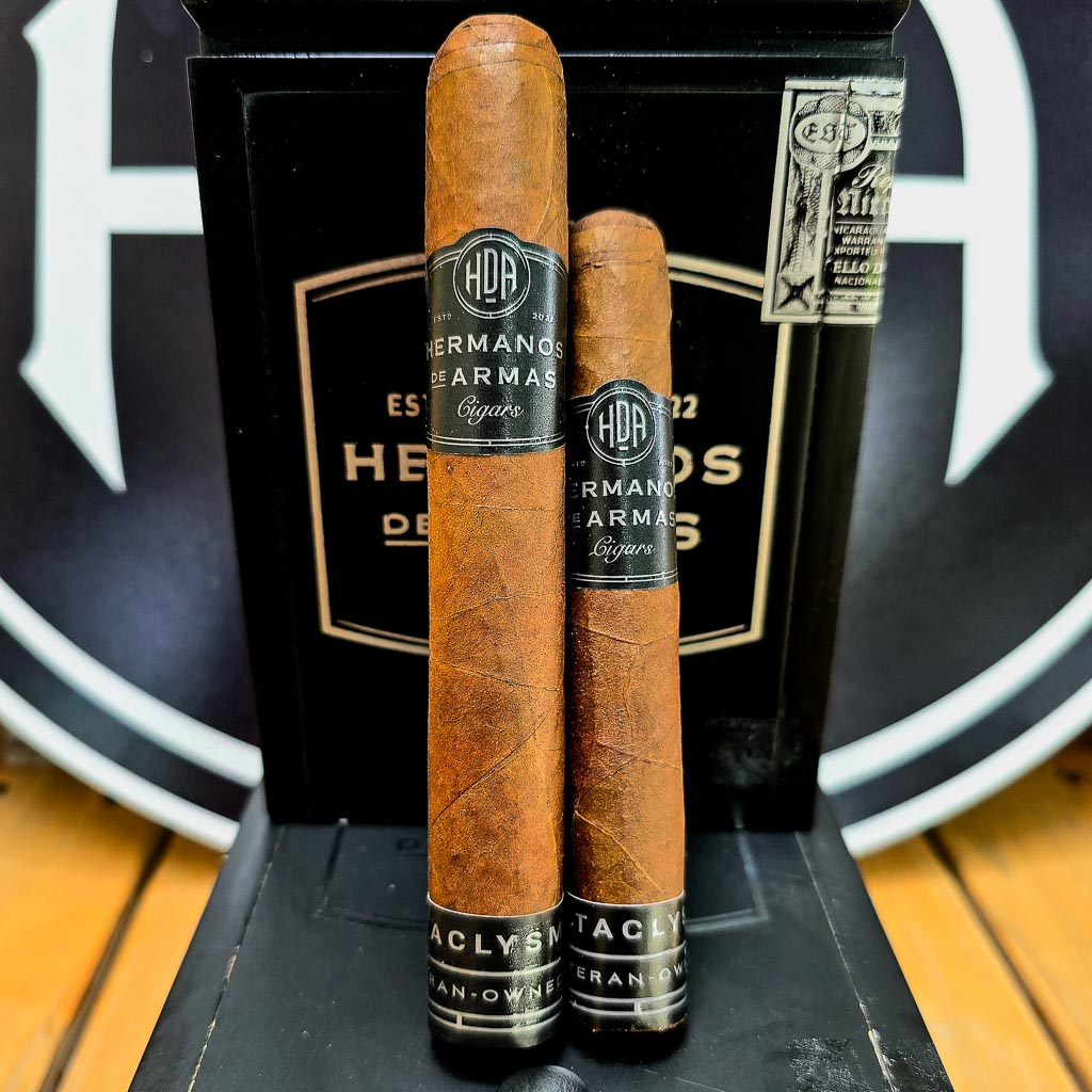 Two Hermanos De Armas Cataclysm cigars standing upright against the black HDA box with bold Hermanos De Armas logo visible in the background at Smokers Abbey Austin