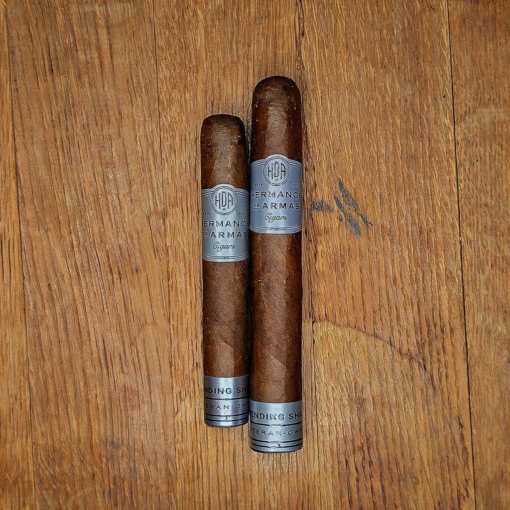 Two Hermanos De Armas Descending Shadows cigars standing upright against the dark gray HDA box with bold black Hermanos De Armas logo visible in the background at Smokers Abbey Austin