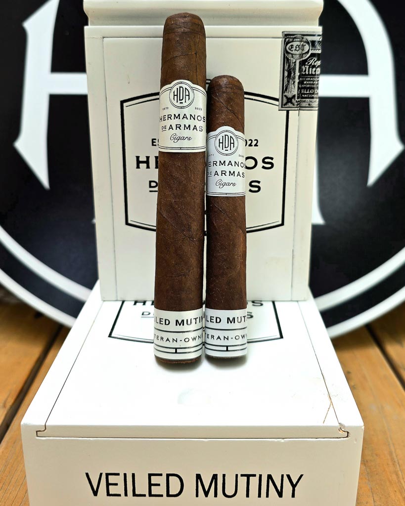 Two Hermanos De Armas Veiled Mutiny cigars standing upright against the white HDA box with bold black Hermanos De Armas logo visible in the background at Smokers Abbey Austin