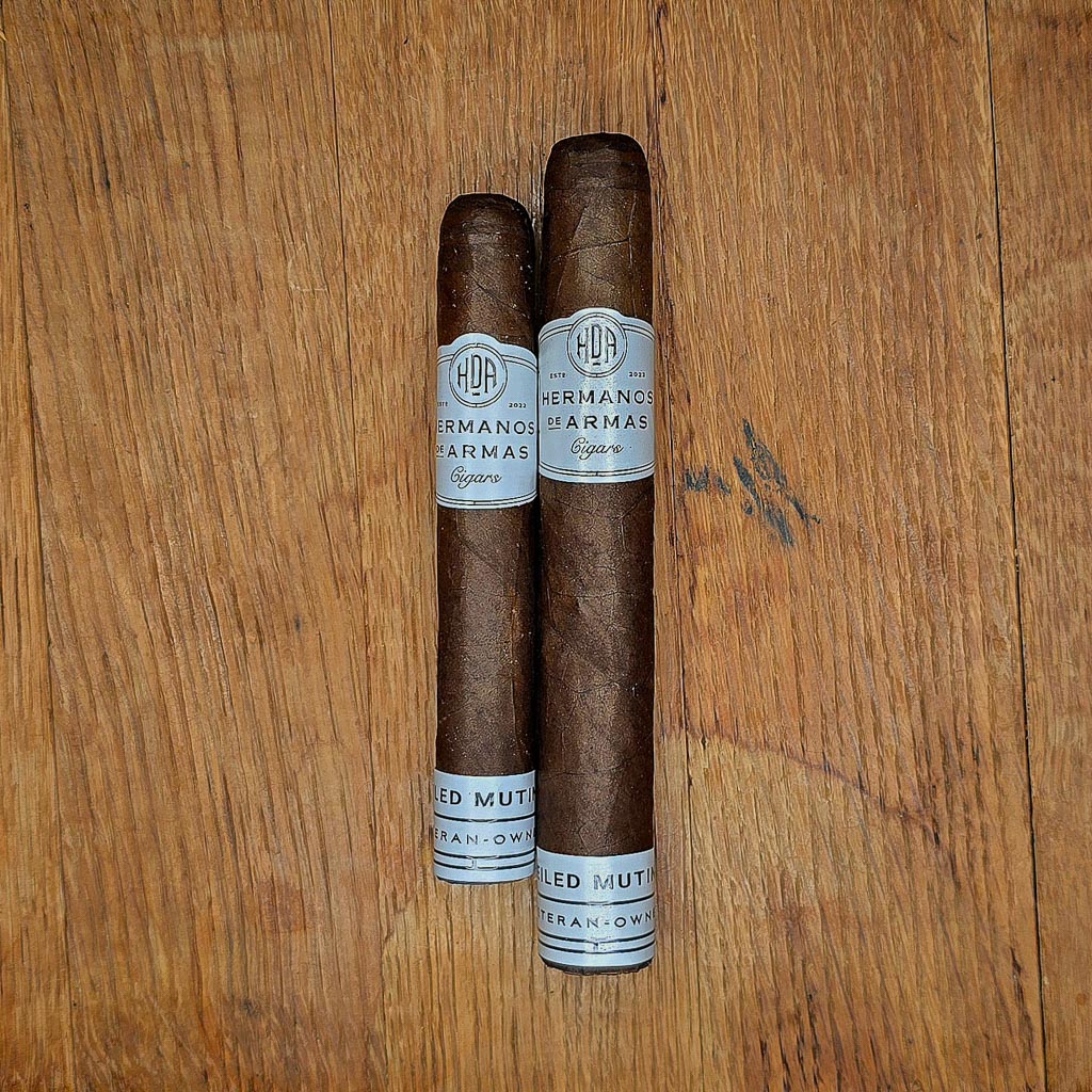 Hermanos De Armas Veiled Mutiny Robusto and Toro cigars displayed side by side on rustic wood surface showing Ecuadorian Maduro wrapper and white HDA band with Veteran-Owned footer at Smokers Abbey Austin