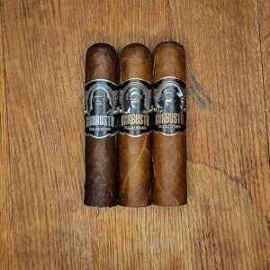 Three JRE Aladino Gorbusto cigars — Gori, Chimp, and Orang — displayed side by side on rustic wood surface showing three distinct wrapper shades at Smokers Abbey Austin