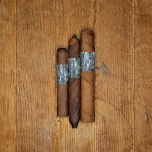 Three La Instructora Box Pressed cigars in No.3, No.4, and No.5 vitolas displayed side by side on rustic wood surface showing three distinct wrapper shades and holographic silver and blue La Instructora band at Smokers Abbey Austin