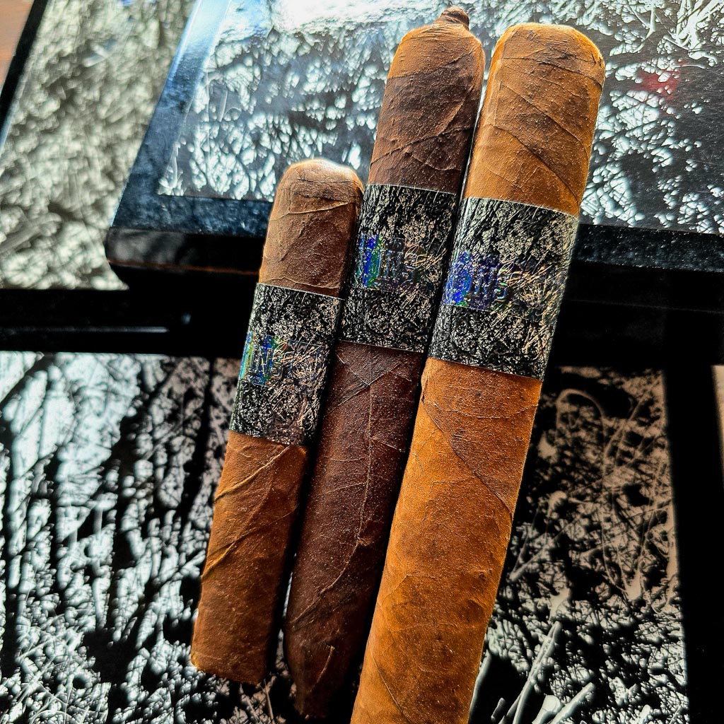 Three La Instructora Box Pressed cigars standing upright against the open black and white La Instructora box with abstract splatter artwork showing Brazilian Cubra and natural wrapper shades at Smokers Abbey Austin.