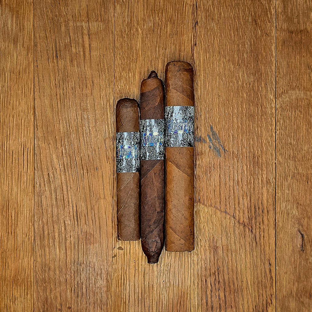 Three La Instructora Box Pressed cigars in No.3, No.4, and No.5 vitolas displayed side by side on rustic wood surface showing three distinct wrapper shades and holographic silver and blue La Instructora band at Smokers Abbey Austin