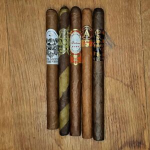 Five premium lancero cigars laid side by side on a wood surface featuring the BLK WKS Santa Muerte, Oscar Valladares 2012 Barberpole, Patina Oro de Nicaragua, JRE Aladino Classic, and Noel Rojas X OSOK Los Tejanos, available in The Lancero Sampler at Smokers Abbey Austin.