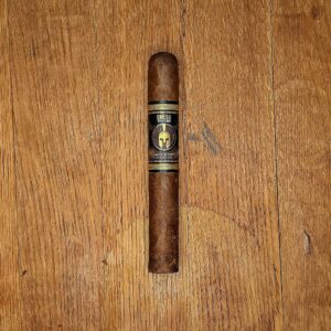 Jake Wyatt Cigar Co. Titan Series Omega Rosado Toro cigar with black and gold Spartan helmet band displayed on rustic wood surface at Smokers Abbey Austin