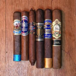 Six curated boutique cigars in the Smokers Abbey Nu New Sampler displayed side by side on rustic wood surface including El Mago Beach Blend, Chateau Diadem Certitude, La Instructora Petite Solomon, Jake Wyatt Titan Series Colossus, E.P. Carrillo Pledge Sojourn, and La Galera 1936 Box Pressed El Lector