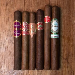 Corona cigar sampler featuring Somm BDX, Matilde Renancer, Artista Puro Ambar, Crowned Heads Four Kicks, Curivari Seleccion Privada, and Patina Sumatra Copper coronas displayed side by side on rustic wood surface at Smokers Abbey Austin