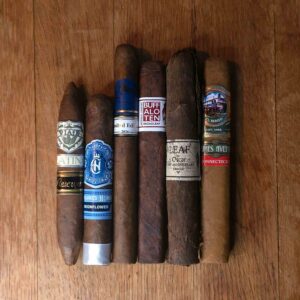 text: Newness Sampler featuring Viaje Platino Reserva, Crowned Heads Moonflower, Cavalier Genève Limited Edition 2026, Artista Buffalo Ten Broadleaf, Oscar Valladares Leaf by Oscar Corojo, and El Mago James Avenue Connecticut displayed side by side on rustic wood surface at Smokers Abbey Austin